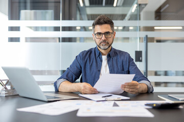 Obraz premium Confident male manager sitting at a desk in a contemporary office, carefully reviewing business documents and financial reports while looking directly at the camera