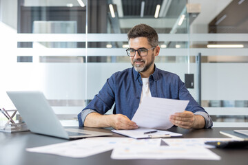 Obraz premium Confident businessman in glasses and denim shirt reads documents at a modern office desk, focused on laptop and paperwork while planning finances and strategy for his startup