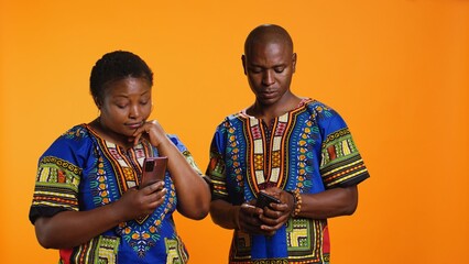 Obraz premium Ethnic people in traditional clothes checking smartphone apps, looking at messages received on social media network. African american man and woman browsing websites on mobile device.