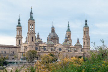 Naklejka premium Majestic Basilica of Our Lady of the Pillar framed by green trees and autumn foliage in Zaragoza, Spain. The historic Spanish cathedral features stunning domes and towers under a soft overcast sky