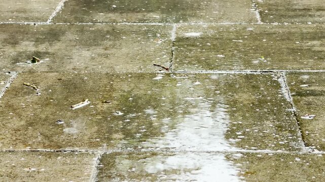 Heavy raindrops splash onto a wet stone walkway outdoors. The paved slabs glisten during a summer shower in Simferopol.