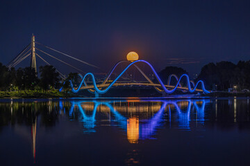 Full moon rises above the illuminated Obolon pedestrian bridge in Kyiv, Ukraine. Color-changing lights reflect on calm water, creating a striking night cityscape and lunar reflection. © elena_suvorova