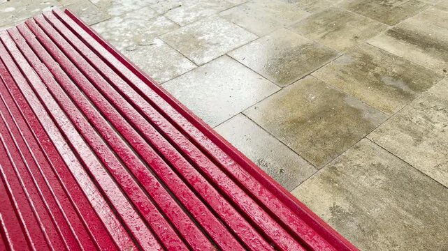 A wet red bench sits empty after a rainstorm. Raindrops glisten on the wooden slats next to damp stone pavement tiles. The public square remains quiet and peaceful.