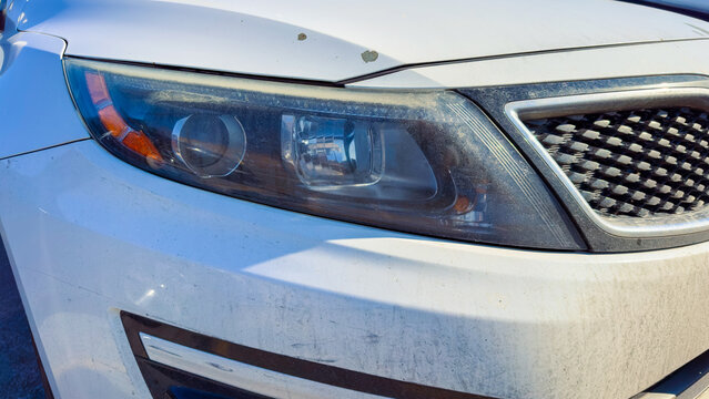 White car parked under sunlight shows details of front headlight and grime indicating lack of recent cleaning