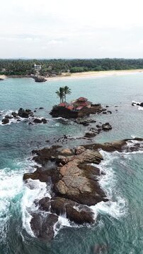 Aerial view of Seenigama Muhudu Dewalaya temple on a rocky island with crashing waves and palm trees near the coast in Peraliya, Sri Lanka.