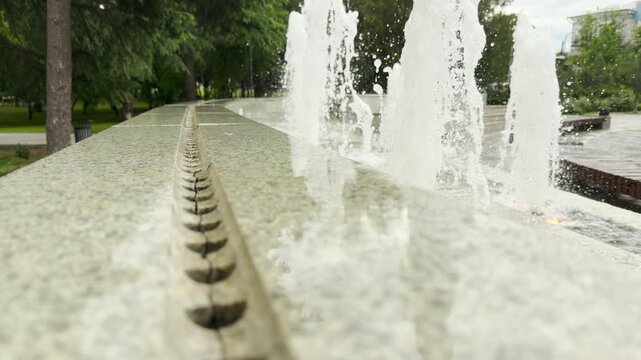White water jets spray upward behind a polished granite ledge. The fountain brings life to a quiet city park. Green trees stand in the background.