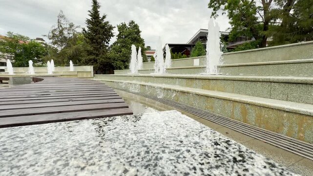 Multiple water jets spray upward from a tiered stone fountain. The water cascades down beside a wooden walkway in a green city park.