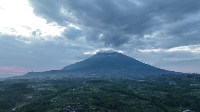 Aerial view of the majestic Mount Merbabu peak shrouded in clouds above lush green terraced agricultural fields at dusk Kecamatan Ngablak, Jawa Tengah, Indonesia.