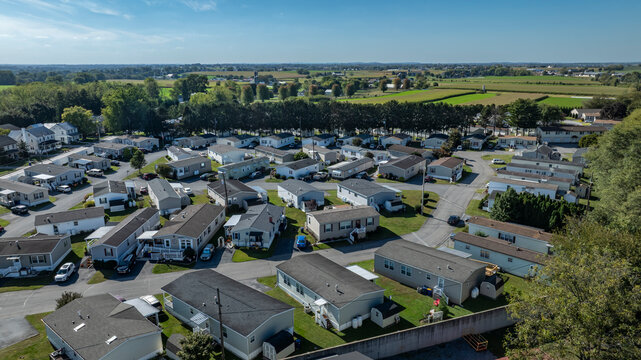People live in a mobile and manufactured home community with rows of houses in a green rural area. The sun shines on the neighborhood and fields surround the homes. Cars are parked along the streets.