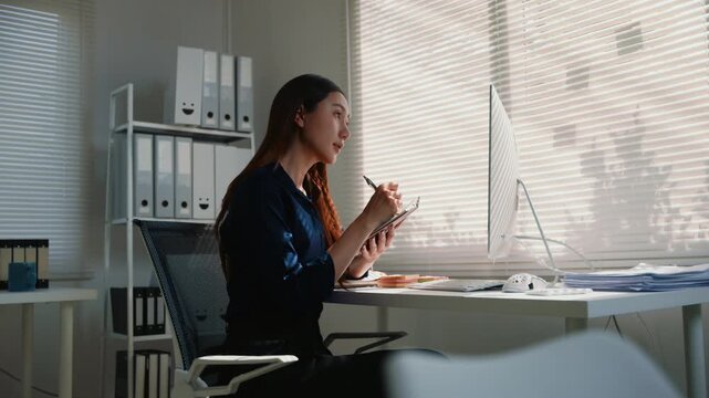 Young Asian business woman sitting at desk using computer and stylus on digital tablet, taking notes during an online meeting