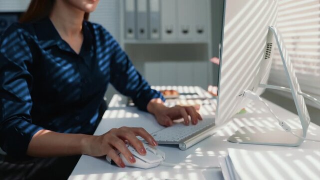 Young businesswoman typing on a computer keyboard and using a mouse while sitting at her desk. Sunlight streams through the blinds