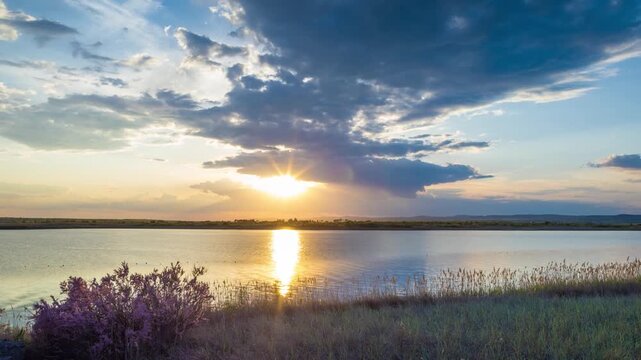 Clouds run across the sky with a sunset light over a lake with herbs on a shore