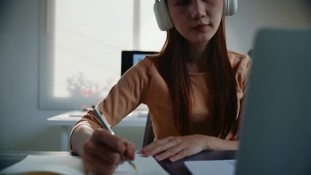 Young woman wearing headphones and taking notes in her notebook while attentively listening to an online class on her laptop at home
