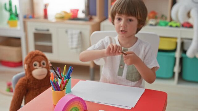 Boy drawing with pencil and paper at small table in a playroom building with stuffed monkey nearby; calm concentration.