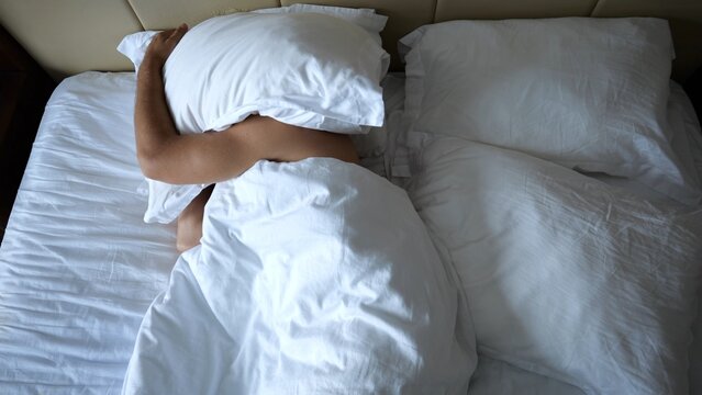 Young man sleeping in large bed and covering head with pillow in modern hotel room or home interior. Guy continuing restful sleep in cozy atmosphere, showing comfort and relaxation. Top view