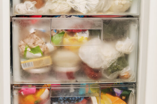 Overfilled freezer drawer packed with frozen food products. Frosted storage compartment in household refrigerator. Food preservation and organization problem background.