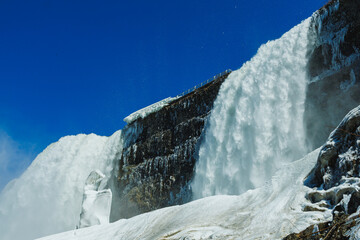 Water flows over Niagara Falls in winter with ice and snow surrounding the rocks and water as it cascades down under a clear blue sky © DSMT