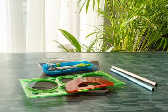 Green geometric ruler, red protractor and compass set in blue case on dark desk with pencils near window with tropical plants. Concept of geometry tools, school supplies and math education
