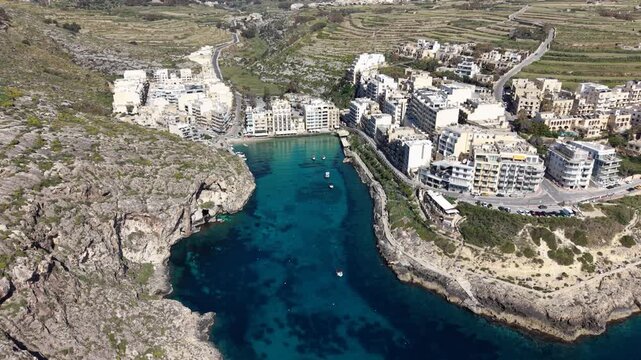 Aerial view of Xlendi Bay featuring turquoise waters, steep limestone cliffs, and coastal buildings under bright sunlight in Munxar, Gozo Region, Malta.