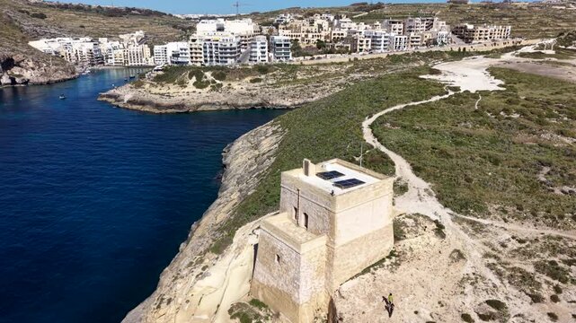 Aerial view of the historic Xlendi Tower standing on a rocky cliff overlooking the blue waters of the bay and the town of Munxar, Gozo Region, Malta.