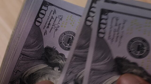 A close-up image of a hand holding a thick stack of one hundred dollar bills. The bills are crisp and layered neatly, showing the portrait of Benjamin Franklin prominently.