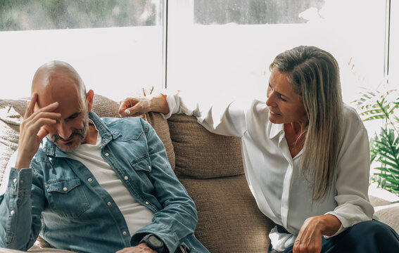 Stressed caucasian man holding head while woman reaches out during tense conversation on couch, depicting relationship conflict, communication problems, and emotional tension