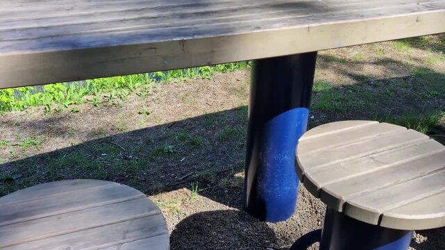 Sunlit circular wooden seats positioned beneath a long wooden table in an outdoor environment. Ground covered partly with dirt and patches of grass, casting both shadows and light.