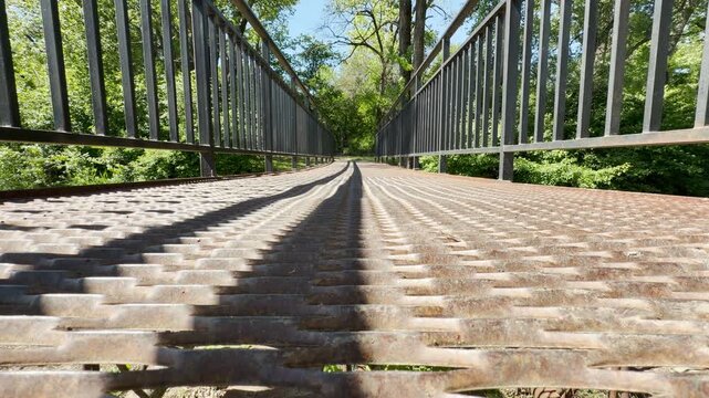 A metal pedestrian bridge extends into a lush, green woodland in a park. The grated walkway features long, parallel shadows cast by the railing. The scene is bright, sunny, and peaceful.