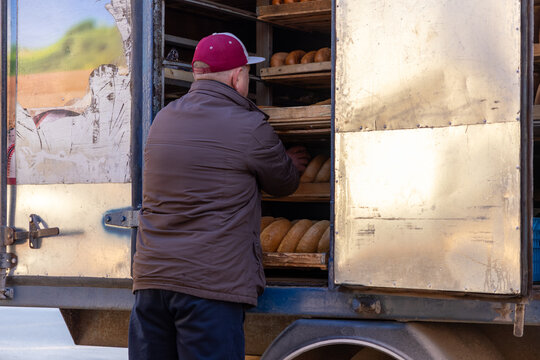 A delivery driver loads fresh bread loaves into metal shelves of a truck, conveying industrial food logistics and wholesale supply chain operations in a daily commercial setting