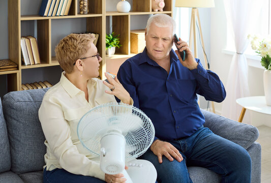 Seniors couple heatwave overheating fan complaint phone repair. At home they call a tech about a failed air conditioner in summer while using a desk fan. Concept shows home cooling crisis.