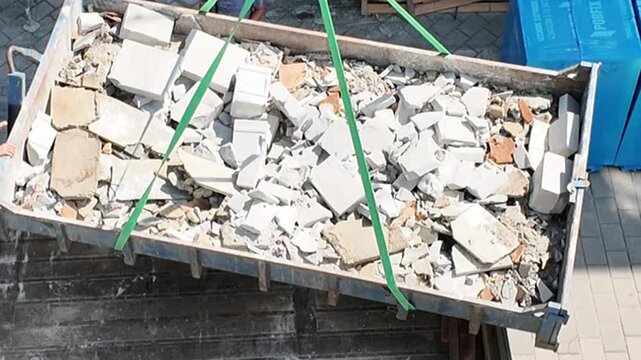 Construction debris being lifted by crane in a skip container at a site. A large skip filled with rubble and construction waste is hoisted by a crane, showcasing waste management processes on a