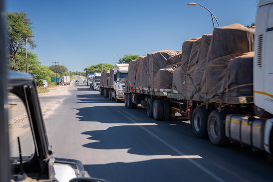 A long line of tarpaulin covered heavy haul trucks moves on a two lane road near the Botswana Zimbabwe border by Chobe National Park under midday sun and deep sky.