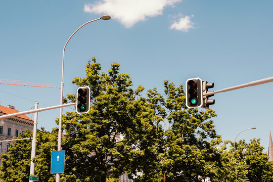 Two traffic signal heads suspended over a tree-lined city street beneath a pale blue sky with scattered clouds. Metal poles and horizontal crossarms hold the