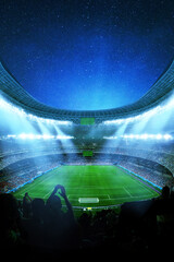 Obraz premium Modern massive modern soccer stadium at night, brightly lit by floodlights. Cheering crowds fill the tiers under a deep blue starry cosmos. Silhouetted fans are in the foreground. Football match