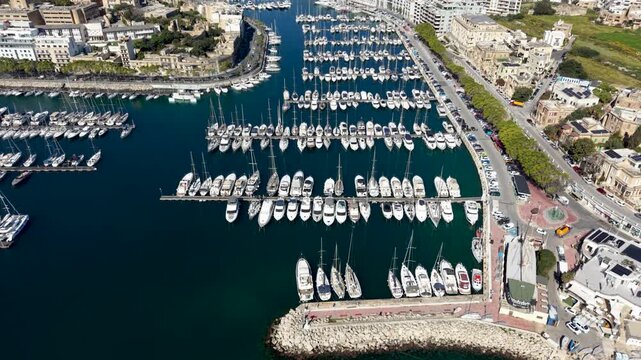 Aerial view of numerous yachts and sailboats docked in neat rows within the marina, featuring the coastal road and urban architecture in Ta' Xbiex, Eastern Region, Malta.