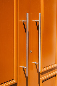Close-up of a painted orange double door featuring two vertical stainless steel pull handles mounted on raised panels. Warm directional lighting casts soft