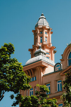 A pale peach historic tower with ornate turrets and a domed roof rises above a tree-lined city street against a clear blue sky. Decorative cornices, arched