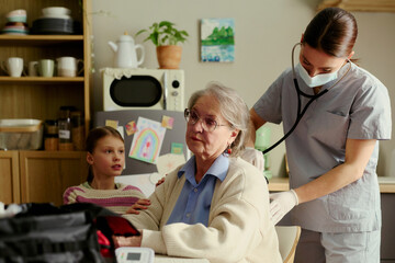 Fototapeta premium Senior woman receiving home care checkup from female nurse while granddaughter supporting nearby, showing eldercare, family support, home healthcare services and caregiving