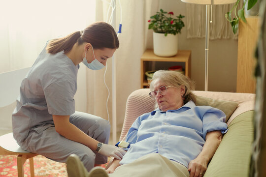 Young adult female caregiver assisting senior woman receiving home care treatment with IV line, supporting eldercare and recovery. Ideal for home healthcare, nursing, caregiving marketing