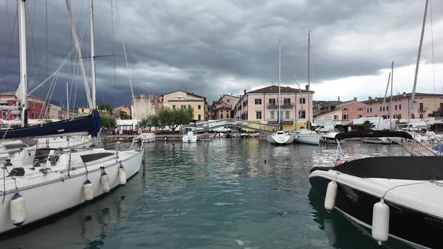 Bardolino, Italy - May 14, 2026 - Sailboats docked in a picturesque harbor with historic buildings under cloudy skies