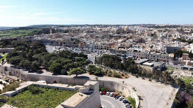 Aerial view of the historic limestone buildings and ancient fortifications of Rabat with green trees and a large parking area under a clear sky in Rabat, Western Region, Malta.