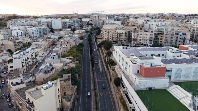 Aerial view of dense urban architecture and a main road passing through the city with a sports field and rooftops under an overcast sky in Msida, Eastern Region, Malta.