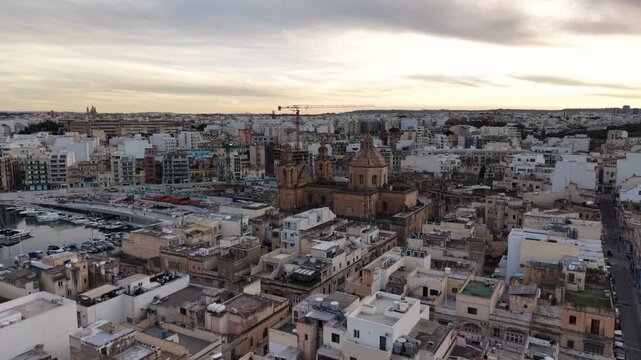 Aerial view of Saint Joseph Parish Church surrounded by dense urban rooftops and the marina with docked boats under a cloudy sky in Msida, Eastern Region, Malta.