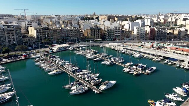 Aerial view of yachts and sailboats docked in the marina surrounded by urban buildings under a clear blue sky in Msida, Eastern Region, Malta.