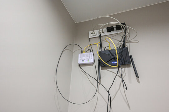 Wall-mounted network setup with black Wi-Fi router, white GPON modem, power strip, tangled Ethernet - fiber cables in corner of light-colored room