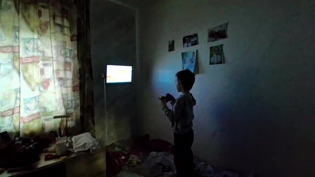 A child is standing in a dark room with a game controller in hand. The TV shows a colorful game while clothes are on the bed. The curtains filter the light from the screen.