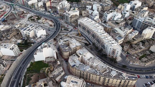 Aerial view of a curved flyover bridge passing through dense urban residential buildings and construction sites in Msida, Eastern Region, Malta.