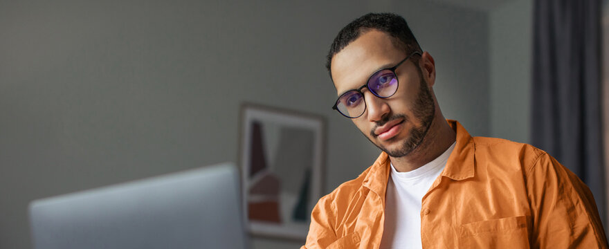 Focused Young Man With Glasses Working On Laptop Computer At Home Office, Sitting At Desk, Engaged In Task. Modern Workspace Reflecting Productivity And Professionalism. Business Concept