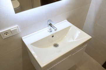Fototapeta premium White ceramic sink mounted on a white vanity unit, paired with a chrome faucet; set against beige tiled walls, with a backlit mirror and electrical outlet nearby