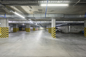 Naklejka premium Spacious underground parking with concrete pillars wrapped in yellow-black hazard stripes, blue overhead pipes, fluorescent lighting, and a few parked cars in marked bays